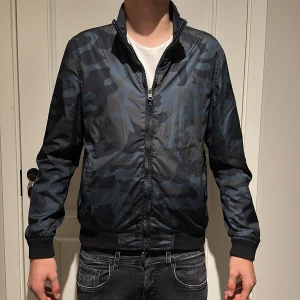 Blå camouflage bomberjacka från Woolrich - Säljer en snygg bomberjacka från Woolrich som är vändbar. Ena sidan är mörkblå med diskret look, andra sidan har blått camouflagemönster. Jackan har dragkedja, ribbade muddar och hög krage. Perfekt för dig som vill kunna variera din stil.