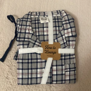 Rutig pyjamasskjorta och byxa från Time to Lounge - Mysig rutig pyjamasskjorta och byxa från Time to Lounge i storlek L. Skjortan har klassisk krage, knappar framtill och ett rutigt mönster i grått, vitt, blått och vinrött. Tillverkad i mjuk bomull som är perfekt för chillkvällar hemma. Oöppnad. 