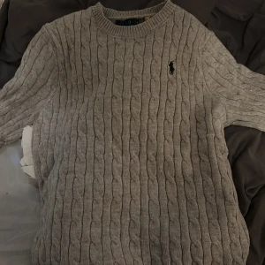 Beige stickad tröja från Polo Ralph Lauren Oanvänd  - Säljer en beige stickad tröja från Polo Ralph Lauren med klassiskt kabelstickat mönster och rund hals. Tröjan har långärmade ribbade muddar och den ikoniska broderade loggan på bröstet. Perfekt för dig som gillar stilren och tidlös design.