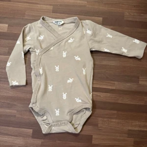 Omlottsbody med kaniner - Mjuk, beige omlottsbody från Kappahl i storlek 62 (2-4mån). Närmast oanvänd utan anmärkningar. Bortryckt tvättråd.
