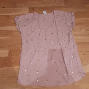 Rosa t-shirt med silverprickar Lindex - Snygg ljusrosa t-shirt från Lindex med korta ärmar och coola silverfärgade prickar över hela framsidan. Tillverkad i mjuk bomull, perfekt för dig som gillar lite extra detaljer på din outfit. Passar dig som vill sticka ut med stil.