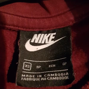 Vinröd Nike hoodie XS - Snygg vinröd hoodie från Nike i storlek XS. Tröjan har huva med snörning, en klassisk magficka och Nike-logga i vitt på bröstet. Perfekt för chill dagar eller när du vill ha en sportig look. Materialet är mjukt och känns skönt mot huden.