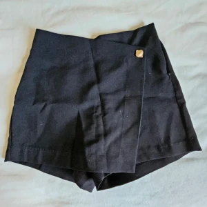 Svarta omlottshorts från Amisu - Snygga svarta shorts från Amisu i omlottmodell med en dekorativ knapp framtill. De är högmidjade och har en stilren, klassisk look som passar perfekt till en trendig outfit. Materialet känns mjukt och lätt, troligen polyesterblandning.