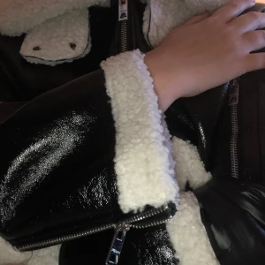 Svart pälsjacka med vita detaljer - Säljer en svart glansig pälsjacka med feta vita teddy-detaljer på krage, ärmslut och fickor. Jackan har dragkedja framtill och coola silvriga detaljer. Perfekt för dig som vill sticka ut och hålla dig varm under kalla dagar.