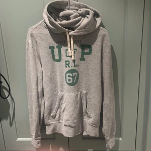 Grå hoodie Polo Ralph Lauren XS - Grå hoodie från Polo Ralph Lauren i storlek XS. Tröjan har grönt tryck med texten 'USGP R.L. 67' på bröstet, snörning vid huvan och en klassisk magficka. Mjuk bomullskänsla och långärmad modell, perfekt för chill dagar.