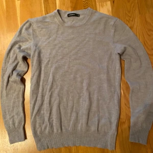 Ljusgrå stickad tröja från Lindbergh 100 merinoull  - En stilren ljusgrå stickad tröja från Lindbergh med rund hals och ribbade muddar vid ärmslut och nederkant. Perfekt för lager-på-lager och passar dig som gillar enkel och clean stil. Materialet är mjukt och lätt, vilket gör den skön att bära.