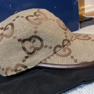 Gucci beige keps med GG-mönster - Snygg beige keps från Gucci med det klassiska GG-mönstret i brunt över hela kepsen. Kepsen har böjd skärm med brun kant och är tillverkad i ett slitstarkt tygmaterial. Perfekt för dig som vill ha en lyxig och trendig accessoar.