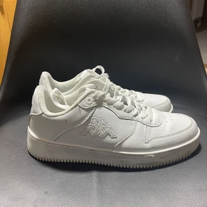 Vita sneakers från Kappa - Fräscha vita sneakers från Kappa med klassisk låg profil och rund tå. Skorna har snörning och Kappas ikoniska logga på sidan. Ovandelen är i syntetmaterial med textilfoder för extra komfort. Perfekta för dig som gillar stilrena och cleana sneakers.