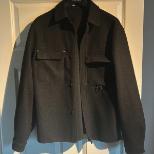 Svart overshirt från Zara i ullmix - Stilren svart overshirt från Zara med klassisk krage, två stora bröstfickor med lock och knappstängning. Jackan är i ullmix och har raka ärmar samt knappar framtill. Perfekt för lager-på-lager och en clean look.