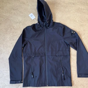 Svart vindjacka från C.P. Company - Svart vindjacka från C.P. Company med huva, dragkedja framtill och två snedställda fickor. Jackan har den ikoniska linsen på ärmen och broderad logga över bröstet. Snygg och stilren design, perfekt för dig som gillar streetwear.