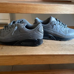 Nike Air Max 90 grå sneakers - Säljer ett par grå Nike Air Max 90 sneakers med svart sula. Skorna har klassisk Air Max-dämpning, snörning och är tillverkade i en mix av syntet och mesh för en sportig look. Perfekta för dig som gillar streetstyle och vill ha bekväma sneakers med ikonisk design.