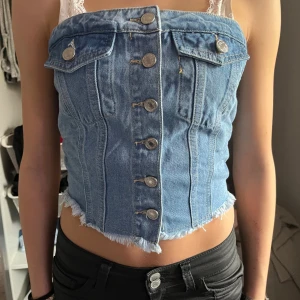 Blå denim tubtopp med knappar - Säljer en blå tubtopp i denim med snygga silverfärgade knappar framtill och fransad nederkant. Toppen har två dekorativa fickor upptill och smockad rygg för skön passform. Perfekt croppad längd och cool streetstil.