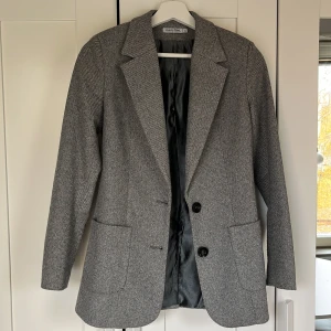 Grå kavaj från Maryline - Snygg grå kavaj från Maryline med diskret randigt mönster. Klassisk passform med två svarta knappar framtill, breda slag och två stora fickor. Fodrad insida och knappdetaljer vid ärmslut. Perfekt för dig som gillar stilrena och tidlösa plagg. 20%Wool 40%Viscose 40% Polyester.