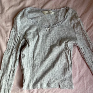 Ljusgrå ribbad topp med spets från H&M - Supersöt ljusgrå långärmad topp från H&M med ribbad struktur och små hålmönster. Rundad halsringning med spetskant och en liten rosett framtill ger en gullig vibe. Perfekt för dig som gillar mjuka material och fina detaljer.