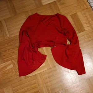 Röd omlottopp med trumpetärmar S - Snygg röd omlottopp med croppad passform och långa trumpetärmar. Toppen har knytband i midjan och är tillverkad i mjuk polyesterblandning. Perfekt för dig som vill sticka ut med en färgstark och trendig look.