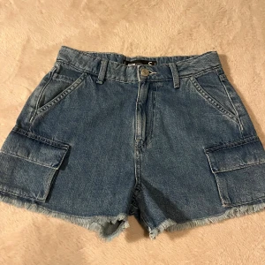 Blå jeansshorts med cargofickor  - Snygga blå jeansshorts från FB sisters köpta på new yorker hög midja, råa kanter och cargofickor på sidorna. Klassisk femficksmodell med bälteshällor och knappstängning. Perfekta för en chill sommarlook.