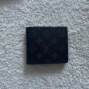 Svart plånbok i skinn - Fin plånbok i svart skinn med klassiskt monogrammönster. Plånboken har flera kortfack, fönster för ID och stilren design. Perfekt storlek för fickan och helt oanvänd.