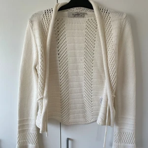 Vit stickad kofta  - Säljer en vit stickad kofta från CLARA i storlek M. Koftan har snygga virkade detaljer, lång ärm och knytband framtill. Perfekt för lager-på-lager och ger en chill men ändå stilren vibe. Materialet känns mjukt och luftigt, passar grymt till både jeans och klänning.
