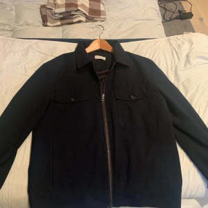 Mörkblå overshirt med dragkedja - Snygg mörkblå overshirt från These Glory Days med dragkedja framtill och två bröstfickor med knappar. Jackan har en klassisk krage, Perfekt för lager-på-lager och passar till jeans eller chinos. Pris är ej hugget i sten, köptes 2023 för 2000kr