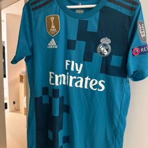 Real Madrid bortatröja Sergio Ramos S - Blå Real Madrid bortatröja från Adidas med Sergio Ramos och nummer 4 på ryggen. Tröjan har korta ärmar, pixelmönster i mörkare blått, klubbmärke, Champions League-patch och Climacool-material. Fly Emirates-logga på bröstet.