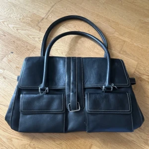 Svart skinnhandväska med fickor - Stilren svart handväska i skinn med två stora ytterfickor framtill och kontrasterande vita sömmar. Väskan har dubbla handtag och flera fack inuti, inklusive ett med dragkedja. Perfekt för dig som vill ha en klassisk och praktisk väska med snygga detaljer.