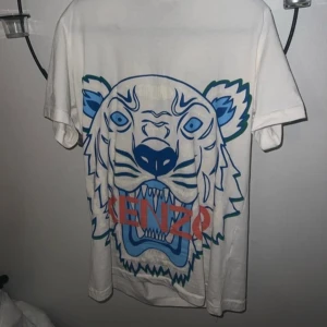 Vit Kenzo t-shirt med tigertryck - Kenzo T-shirt – Vit med exklusivt tigertryck  Säljer en vit Kenzo T-shirt med det ikoniska tigertrycket i blått och grönt samt en röd Kenzo-logga framtill. Tillverkad i mjukt, lyxigt bomullstyg med perfekt passform och rund hals.  En tidlös designklassiker som andas stil, kvalitet och självsäkerhet – perfekt för dig som vill lyfta varje outfit med en unik och exklusiv look.