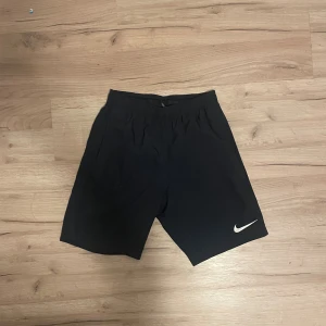 Mörkblå Nike Dri-Fit träningsshorts - Svarta träningsshorts från Nike med Dri-Fit-teknologi som håller dig torr och bekväm. De har elastisk midja och en stilren design med vit Nike-logga på benet. Perfekta för gymmet eller sport, tillverkade i lätt polyester för maximal rörelsefrihet.
