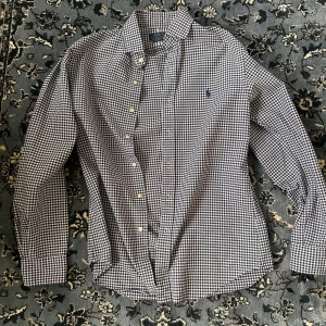 Rutig skjorta från Polo Ralph Lauren - Snygg rutig skjorta från Polo Ralph Lauren i klassisk blå och vit färg. Skjortan har lång ärm, knappar framtill och den ikoniska broderade loggan på bröstet. Perfekt för dig som gillar stilrena och tidlösa plagg med en preppy vibe.
