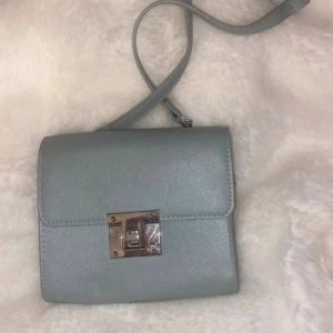 Ljusblå axelväska med silverdetaljer - Snygg ljusblå axelväska i stilren design med justerbar rem och silverfärgat lås framtill. Väskan är tillverkad i ett slätt syntetmaterial och har en kedjedetalj på remmen som ger en cool touch. Perfekt storlek för det viktigaste och passar till många outfits.