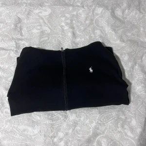 Svart hoodie från Polo Ralph Lauren - Svart hoodie från Polo Ralph Lauren med dragkedja framtill, huva med snörning och två fickor. Klassisk vit broderad logga på bröstet. Skön och stilren modell i mjuk bomull, perfekt för chill dagar eller när du vill ha en clean look. Säljer eftersom den ej passar mig längre.