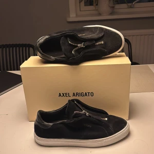Svarta sneakers Axel Arigato mocka - Svarta sneakers från Axel Arigato i mjuk mocka med vit platt sula. Skorna har en unik dragkedjedetalj på ovansidan istället för klassisk snörning, vilket ger en clean och modern look. Perfekta för dig som gillar stilrena och trendiga skor.
