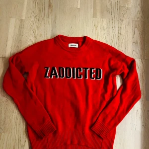 Röd stickad tröja Zadig & Voltaire - Cool röd stickad tröja från Zadig & Voltaire med texten 'ZADDICTED' broderad i svart och vitt på bröstet. Rund halsringning och långa ärmar med ribbade muddar. Perfekt för dig som gillar statement-plagg och vill sticka ut.