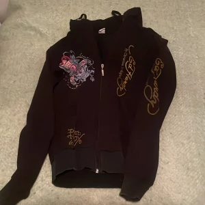 Svart Ed Hardy hoodie med tryck - Säljer en svart hoodie från Ed Hardy i storlek XL  men passar som s med ikoniskt färgglatt tattoo-inspirerat tryck på både fram- och baksida. Tröjan har huva, dragkedja och guldiga loggor på ärmarna. Perfekt för dig som gillar streetstyle och edgy detaljer.pris kan diskuteras. 