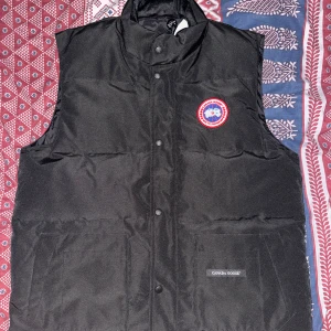 Svart dunväst från Canada Goose - Svart dunväst från Canada Goose med klassisk logga på bröstet och diskret varumärkesdetalj på fickan. Västen har hög krage, dragkedja och tryckknappar framtill. Perfekt för lager-på-lager och riktigt snygg till streetwear.