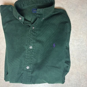 Grön manchesterskjorta Ralph Lauren - Mörkgrön manchesterskjorta från Ralph Lauren med klassisk krage, knappar framtill och lila broderad logga på bröstet. Skjortan har lång ärm och relaxed passform, perfekt för en chill och stilren look.