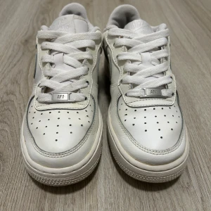 Nike Air Force 1 vita sneakers - Klassiska Nike Air Force 1 sneakers i helvitt skinn med perforerad tå, rund tå och platt sula. Swoosh-logga på sidan och AIR-detalj på sulan. Lågt skaft och snörning framtill. Perfekt för dig som gillar clean och tidlös streetstil.