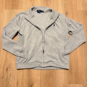 Grå zip-kofta från Polo Ralph Lauren - Snygg grå kofta från Polo Ralph Lauren med klassisk blå logga broderad på bröstet. Modellen har lång ärm, dragkedja framtill och en mjuk bomullskänsla. Perfekt för lager-på-lager och chill dagar. Custom fit för en schysst passform. Jag skulle säga om du har S den här skulle passa dig eftersom det är custom fit