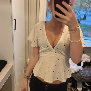 Vit prickig blus med knyt i midjan - Supersöt vit blus med svarta prickar och djup v-ringning. Blusen har korta, lite fladdriga ärmar och knytband i midjan som ger en snygg siluett. Tillverkad i mjuk bomull, perfekt för dig som gillar en feminin och trendig look.
