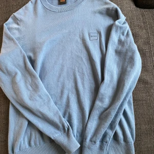 Ljusblå stickad tröja från BOSS - Snygg ljusblå stickad tröja från BOSS med diskret logga på bröstet. Tröjan har rund halsringning, ribbade muddar och är långärmad. Perfekt för dig som gillar stilrena och klassiska plagg med en modern touch.