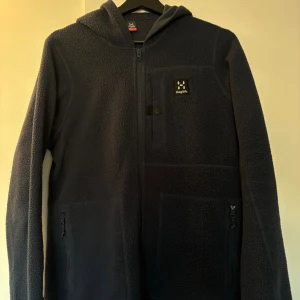 Mörkblå fleecejacka från Haglöfs - Mysig mörkblå fleecejacka från Haglöfs med huva och hel dragkedja. Jackan har två sidofickor med dragkedja och en bröstficka med knappdetalj. Perfekt för kyliga dagar och outdoor-häng.