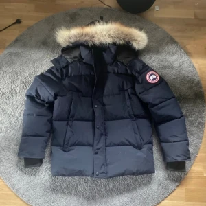 Mörkblå dunjacka från Canada Goose - Säljer en mörkblå dunjacka från Canada Goose med ikonisk patch på ärmen. Jackan har en stor huva med äkta päls, svarta detaljer vid axlarna och ribbade muddar. Fodret är svart och jackan stängs med både dragkedja och knappar. Perfekt för kalla vinterdagar.