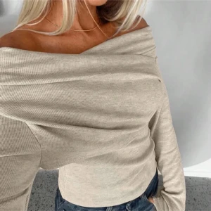 Beige offshoulder topp - Snygg beige offshoulder topp med ribbad struktur och lång ärm. Toppen har en draperad halsringning som ger en cool och modern look. Perfekt att styla med jeans eller kjol för en trendig outfit. Materialet känns mjukt och stretchigt mot huden.