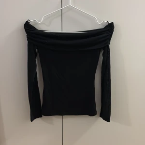 Svart offshoulder topp - Snygg svart offshoulder topp med långa ärmar och bred, draperad kant upptill. Perfekt för dig som vill ha en stilren och trendig look. Toppens passform är tight och materialet känns mjukt mot huden.