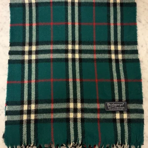Rutig grön halsduk från Burberrys - Snygg halsduk från Burberrys i klassiskt rutigt mönster med gröna, svarta, vita och röda toner. Gjord av 100% lammull och har tvinnade fransar i ändarna. Perfekt för att ge din outfit en cool och tidlös touch.