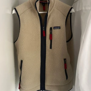 Beige fleeceväst från Patagonia (BYTE) - Säljer en beige fleeceväst från Patagonia i storlek S. Västen har marinblå detaljer, tre fickor med dragkedja och röda zip-pullers. Perfekt för lager-på-lager och har en mjuk, fluffig känsla. Klassisk design med hög krage och diskret logga på bröstet.  (Kan tänkas byta mot mörkblå Patagonia väst )