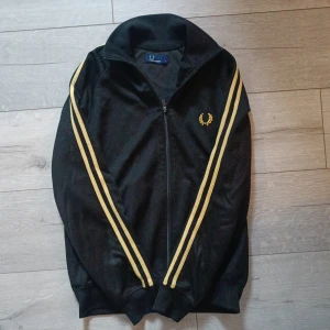 Svart Fred Perry hoodie med gula ränder - Svart hoodie från Fred Perry med två gula ränder längs ärmarna och gul logga på bröstet. Tröjan har dragkedja framtill, ribbade muddar och hög krage. Perfekt för dig som gillar sportig och retro stil.
