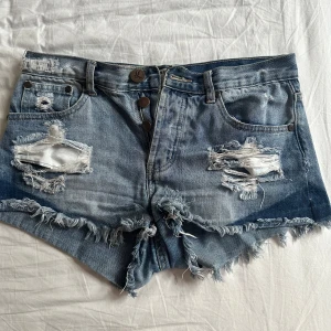 Blå slitna jeansshorts One Teaspoon - Riktigt snygga blå jeansshorts från One Teaspoon med rejält slitna detaljer, fransig kant och klassisk femficksmodell. Shortsen har hög midja och är perfekta för en avslappnad och trendig look. Tillverkade i bomull för skön känsla.