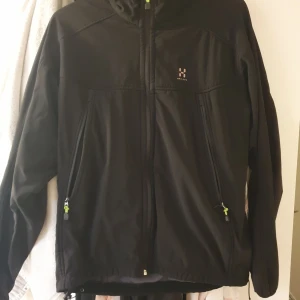 Svart vindjacka från Haglöfs - Svart vindjacka från Haglöfs med hel dragkedja och två stora fickor framtill med gröna detaljer på dragkedjorna. Jackan har hög krage och är tillverkad i ett lätt syntetmaterial, perfekt för blåsiga dagar. Klassisk och stilren design med diskret logga på bröstet. Dock är dragkedjan på ena fickan sönder. 