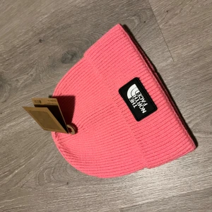 Rosa mössa från The North Face - Snygg rosa ribbstickad mössa från The North Face med klassisk svart och vit logotyp framtill. Mössan har uppvikt kant och är tillverkad i mjukt, stickat material som håller dig varm och ger en cool streetstyle-vibe.