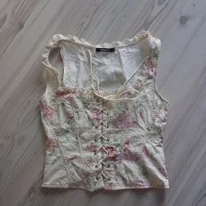 Korsetttopp🦋🌸🌺🌹🪷 - Romantisk korsettopp från Gina Tricot i ljusbeige med blommigt mönster i rosa och grönt. Toppen har snörning framtill, dragkedja i sidan och spetsdetaljer längs halsringningen. Perfekt för dig som gillar vintage vibes och söta detaljer.